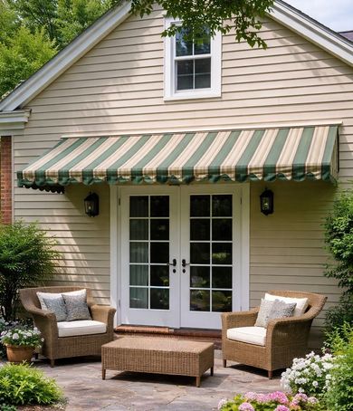 A patio scene with two wicker chairs and a matching coffee table under a green and beige striped awning against a house.