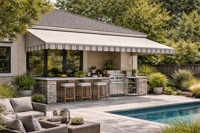 A stone-covered outdoor kitchen with bar stools by a pool under a white awning, surrounded by lush greenery.