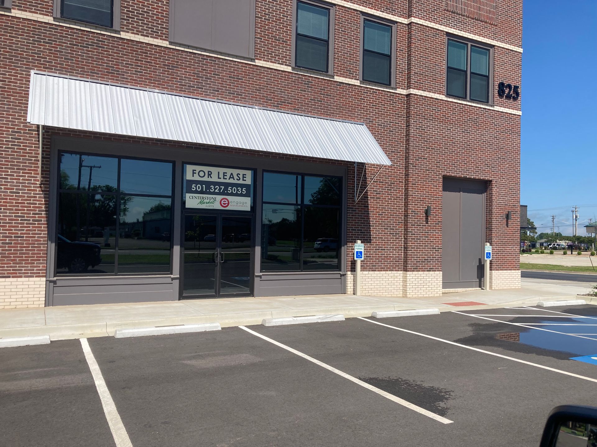 A brick building with a For Lease sign in the ground floor window, featuring a metal awning and a paved parking lot.