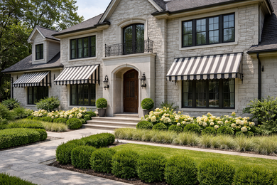 A stone two-story home with striped awnings over the windows and a neatly landscaped front yard with green shrubs.