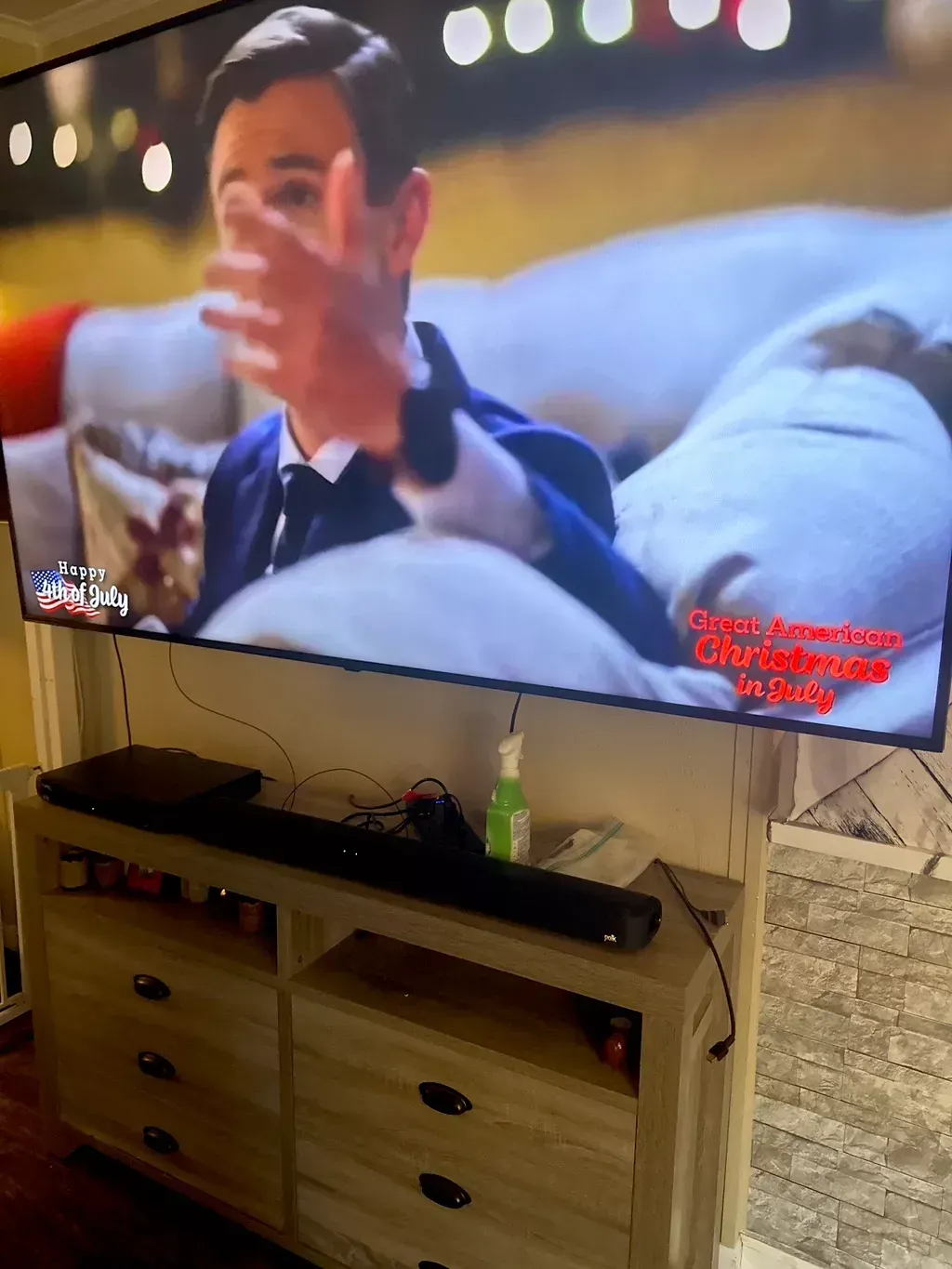 A man on a TV is gesturing; a dresser with a soundbar and electronics is below.