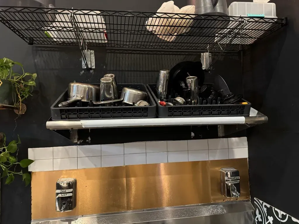 Dishes drying above a sink. Metal shelves hold trays of pots, glasses, and plates. A copper-colored backsplash.