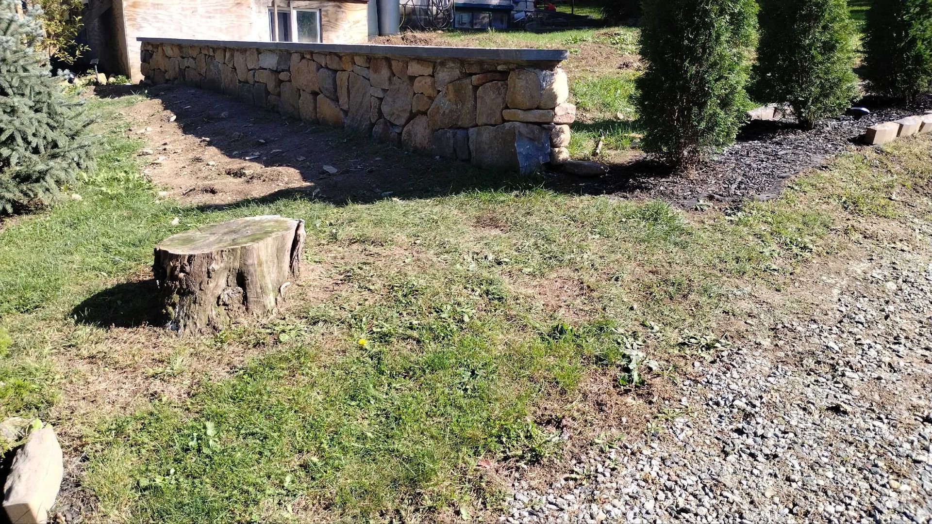 Stone wall with a stump in front, grass, and a gravel path.