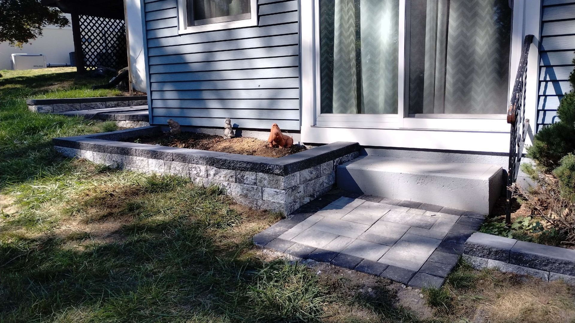 Gray-sided building with low stone planter boxes, concrete steps, and a brick pathway leading to a sliding door.