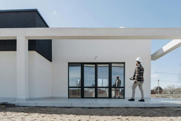Man in hard hat walks past a white building with black trim, holding plans.