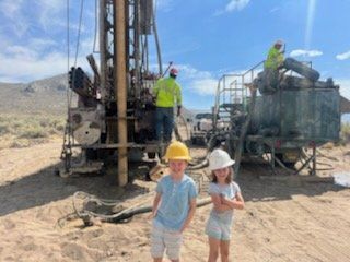 Two children in hard hats stand in front of a drilling rig, with workers in the background. Sunny, outdoor setting.
