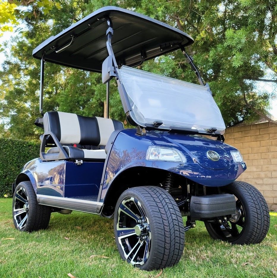 A blue golf cart is parked in the grass in front of a brick wall.