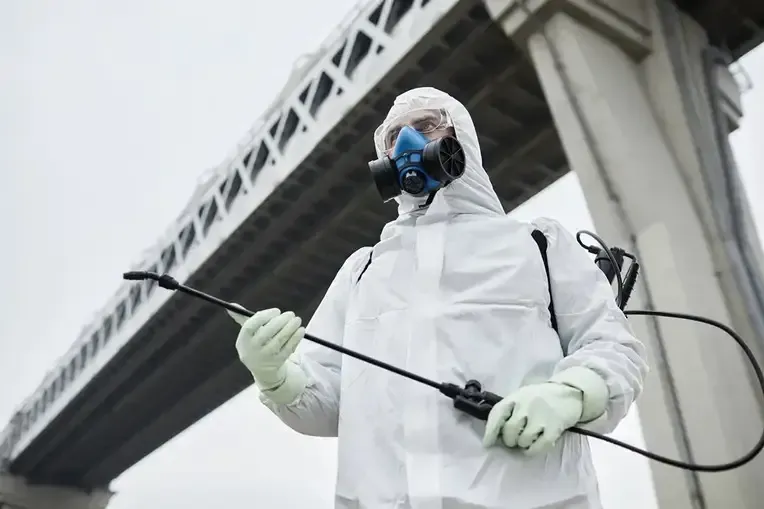 A Man in a Protective Suit and Mask is Spraying a Bridge With a Sprayer — TCB Pest Control Canberra In Greenway, ACT