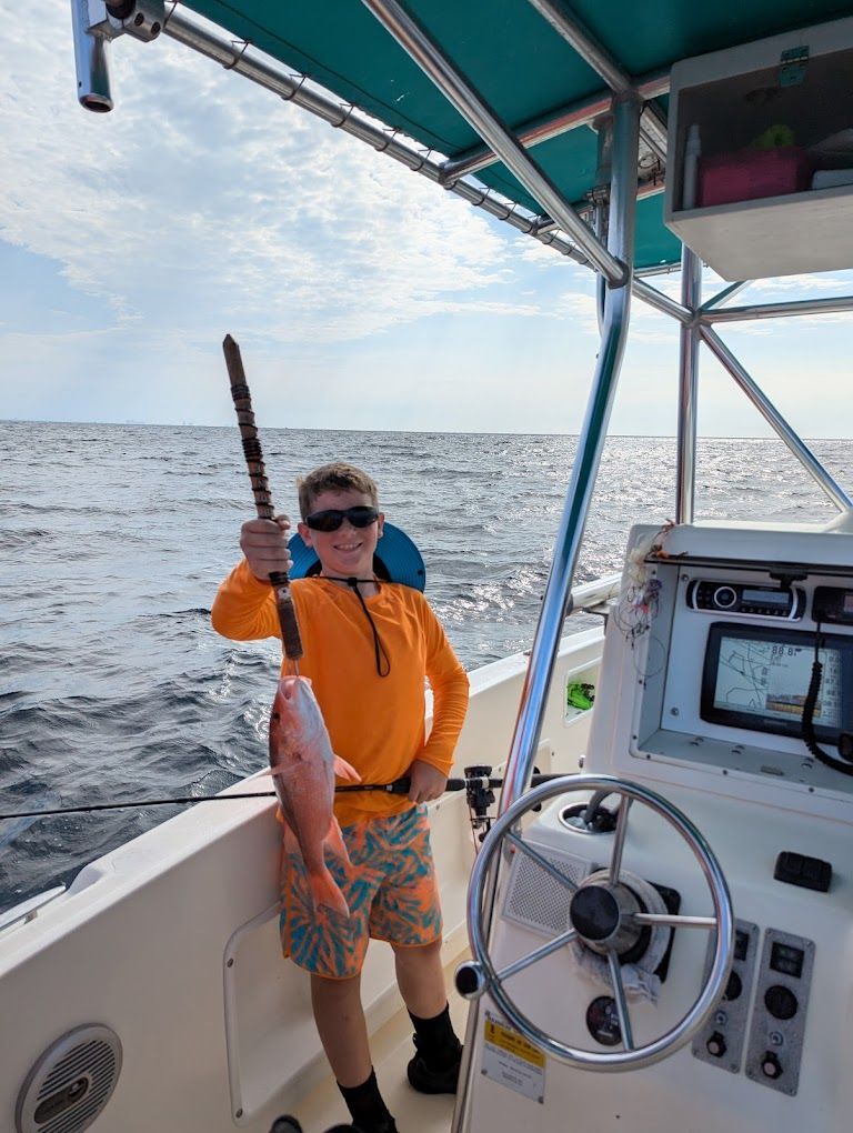 A young boy is standing on a boat holding a fish and a fishing rod.