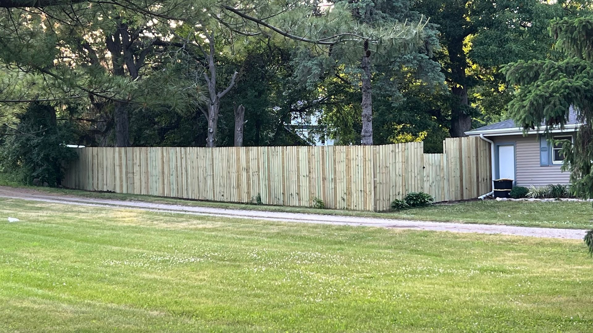 A wooden fence is surrounded by trees and grass in front of a house.