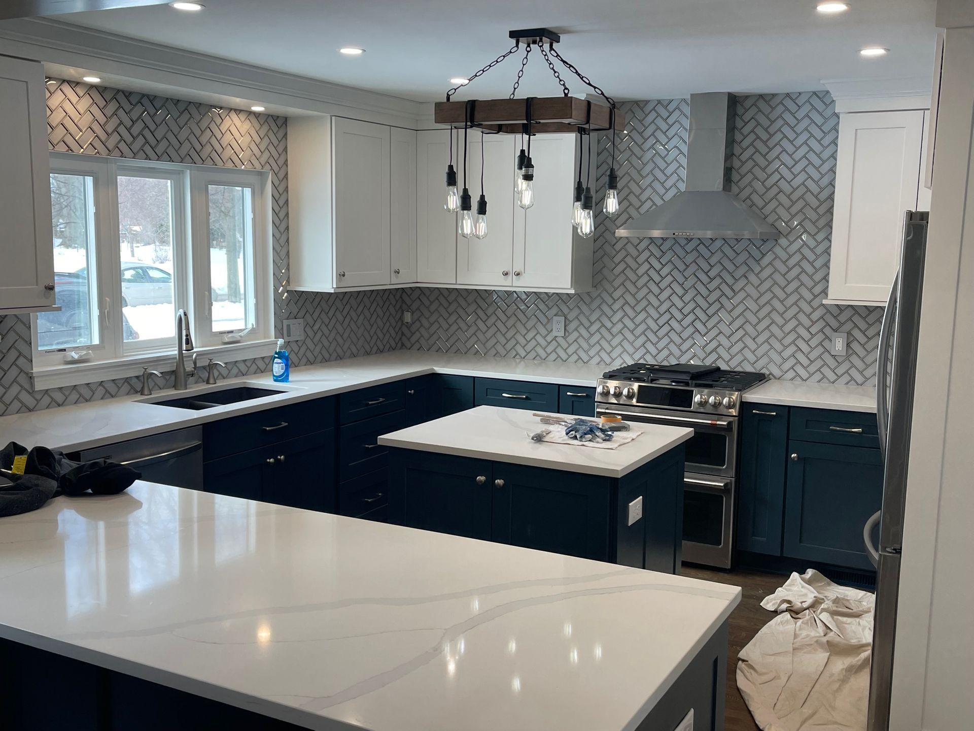 Modern kitchen renovation with navy blue and white cabinets, white countertops, and gray herringbone backsplash.