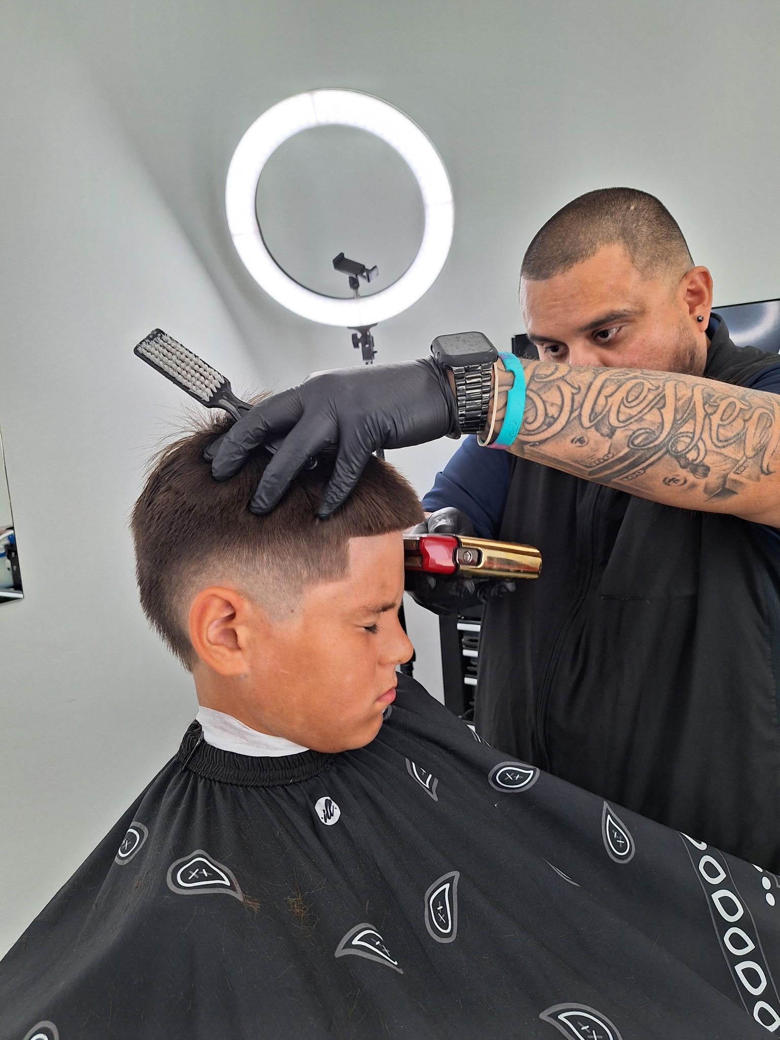 Barber giving a boy a haircut, using clippers, indoors with ring light. Boy wearing cape.