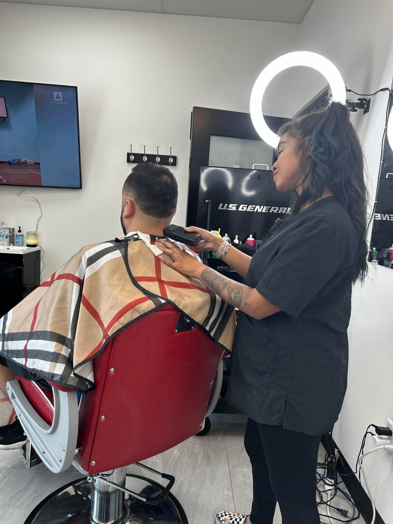 Barber giving a client a haircut in a shop; client in a chair, wearing a cape; barber using clippers.