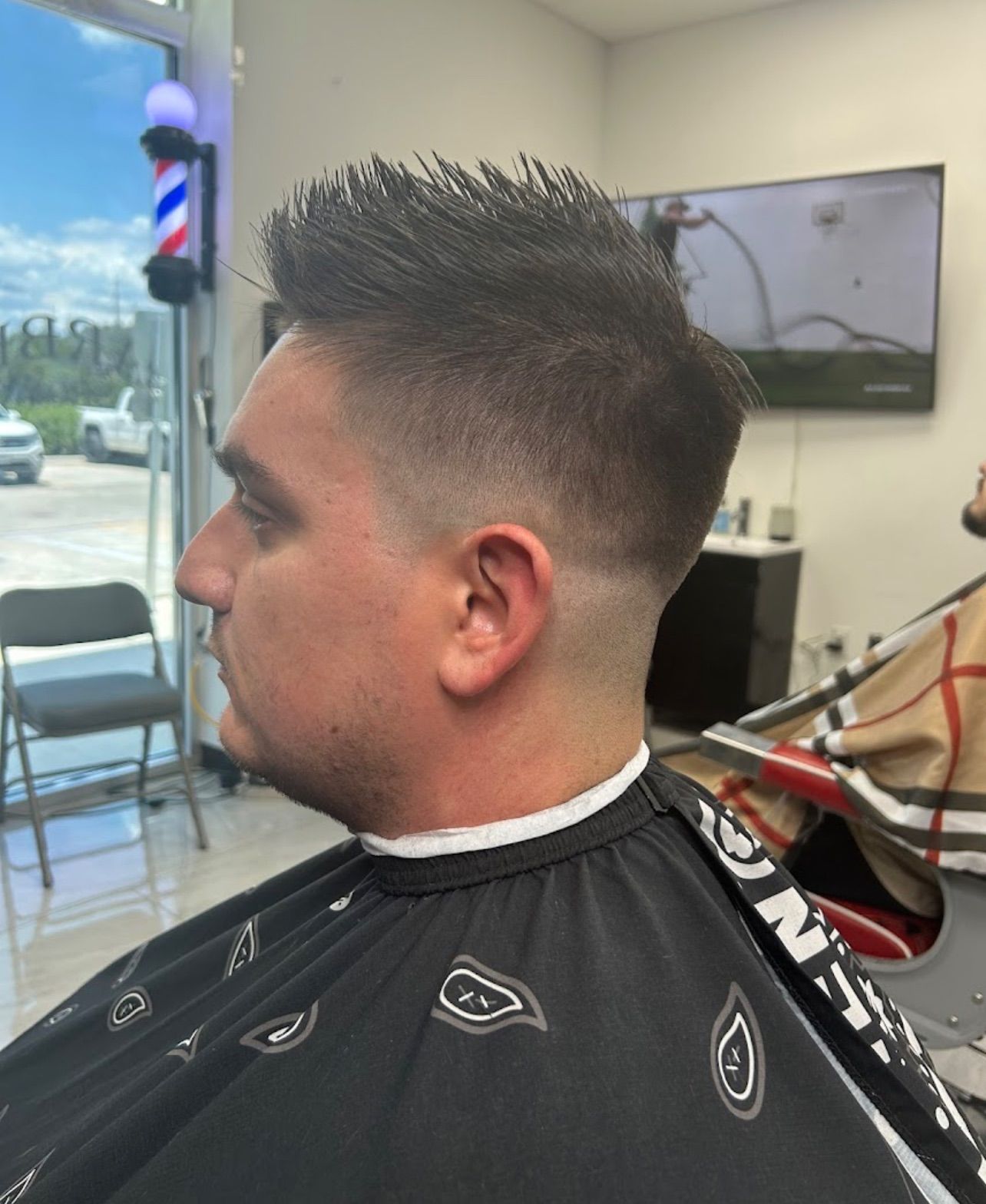 Man getting a haircut in a barber shop. Faded sides, spiky top, blue/white barber pole visible.