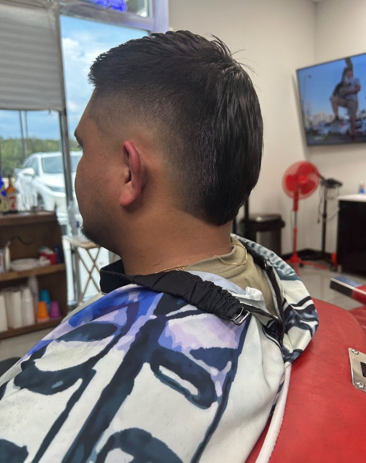Man in a barber's chair with a faded mullet haircut. Wearing a patterned cape, in a barbershop setting.