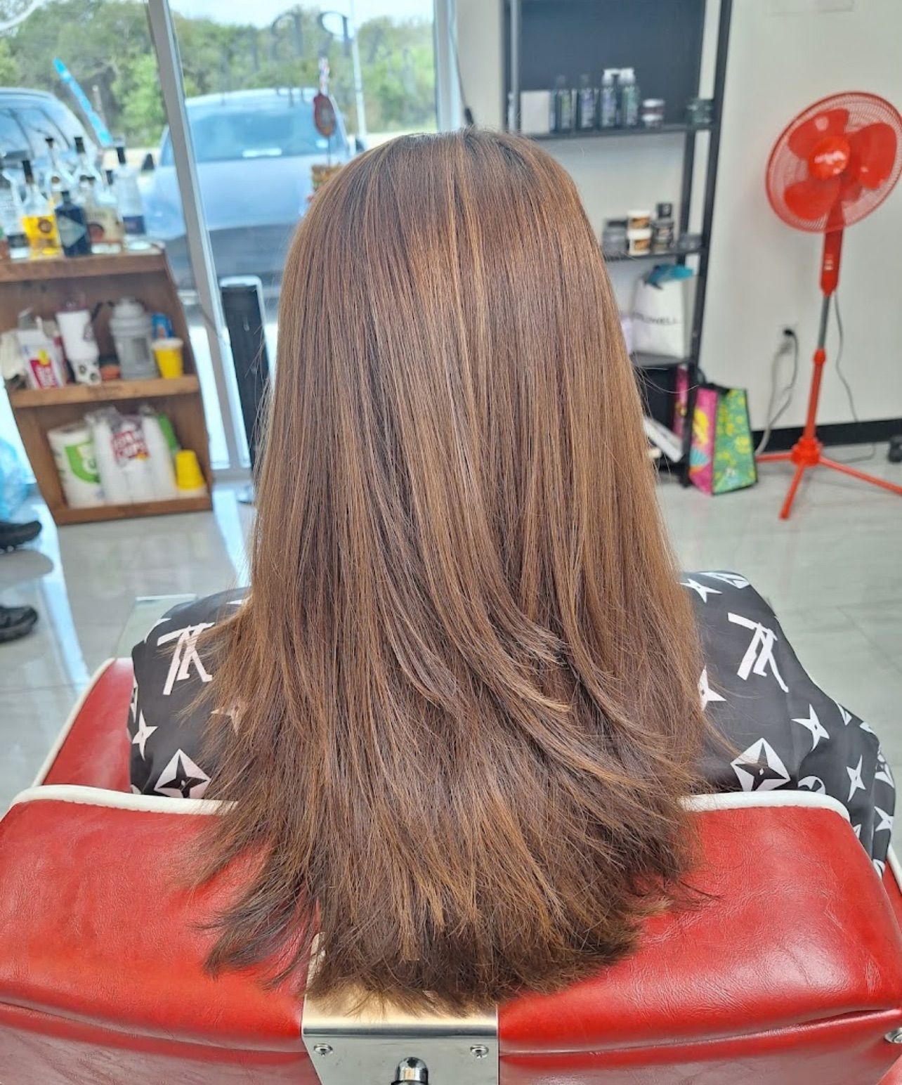 Woman with layered brown hair sitting in a salon chair; red chair, fan in the background.