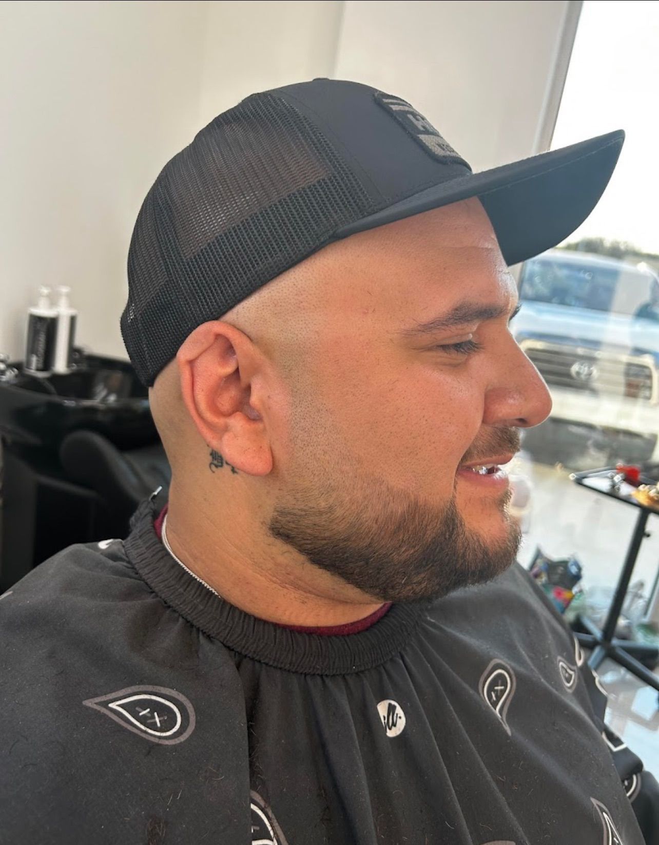 Man with a beard, wearing a black cap and barber's cape, getting a haircut.