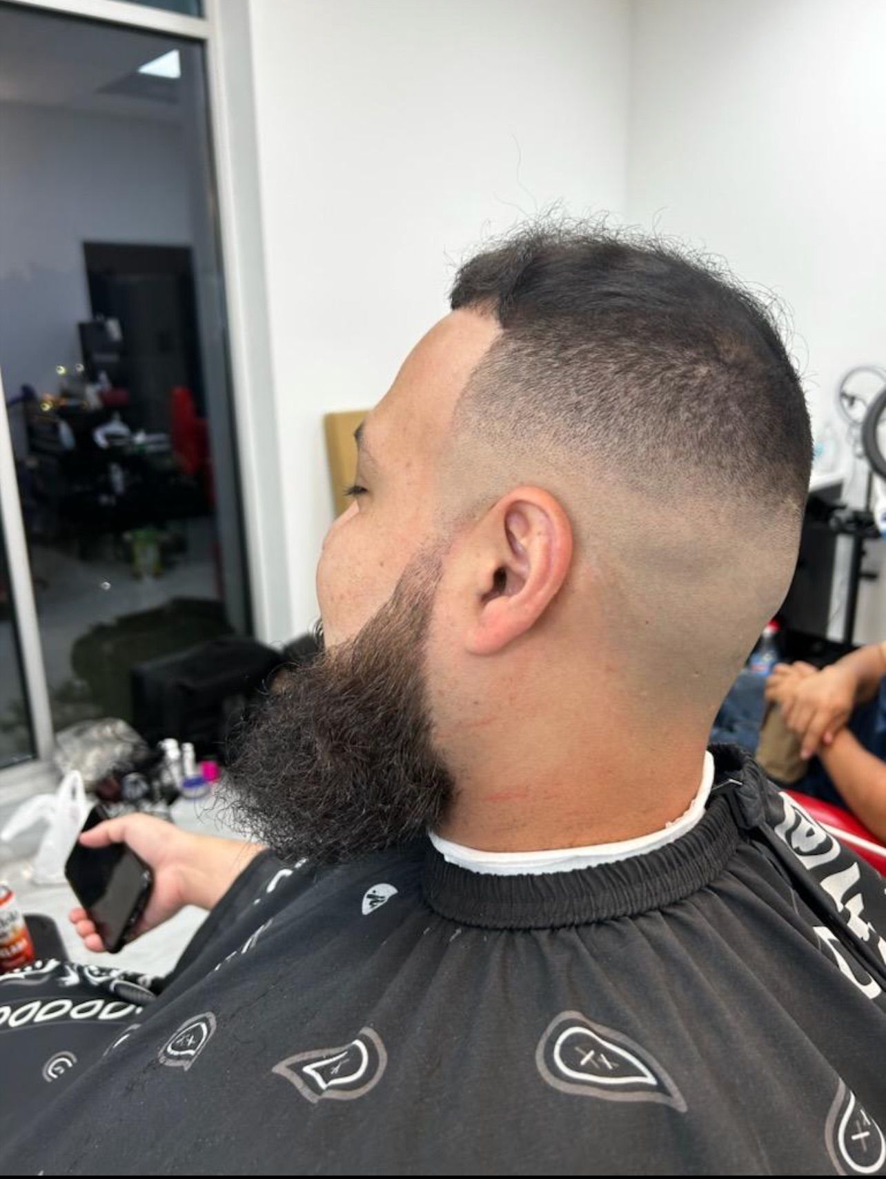 Man with a tapered haircut and beard holding a phone in a barbershop.