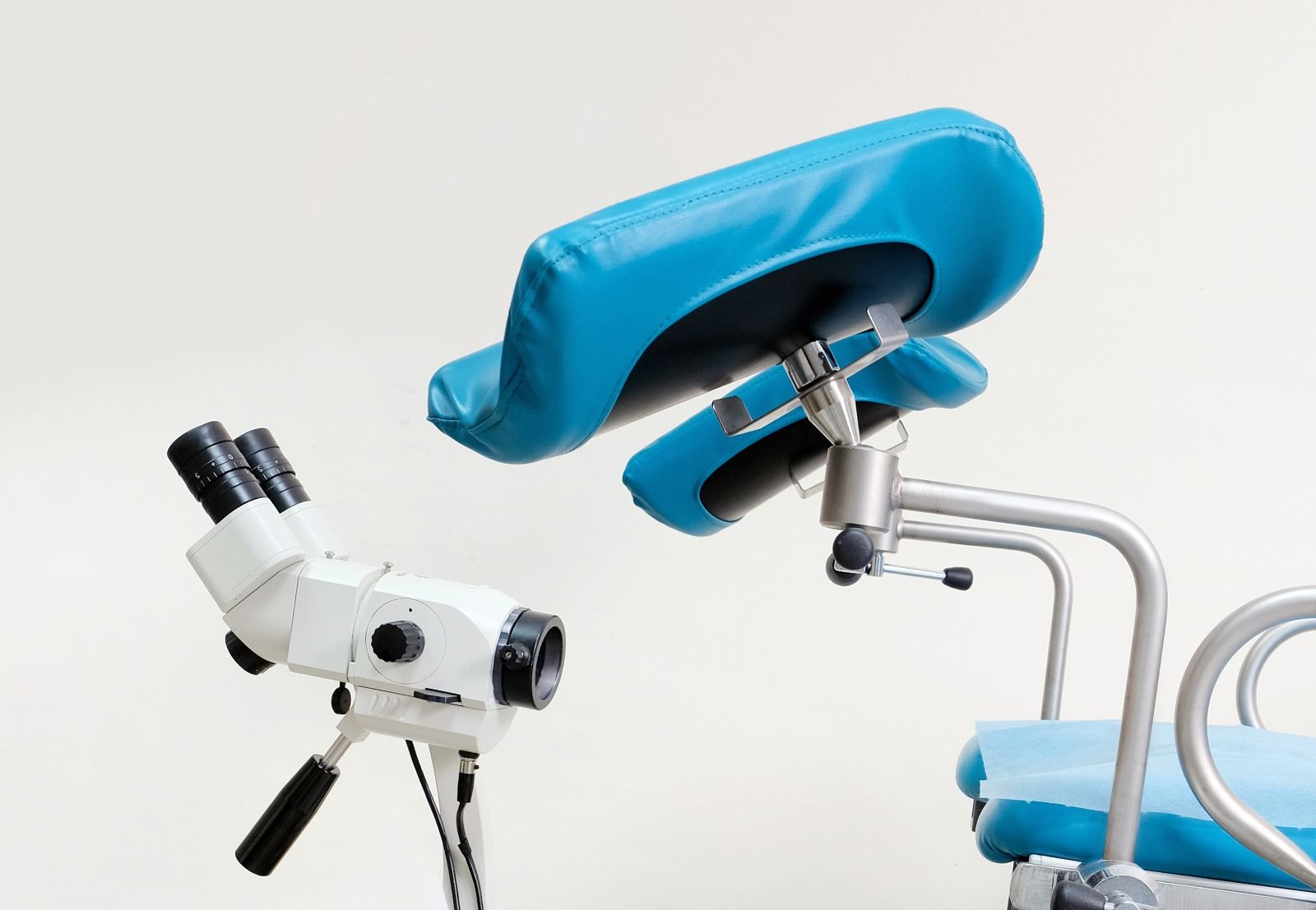 Colposcopio y silla de exploración en un entorno médico, con secciones acolchadas de color azul y fondo blanco.