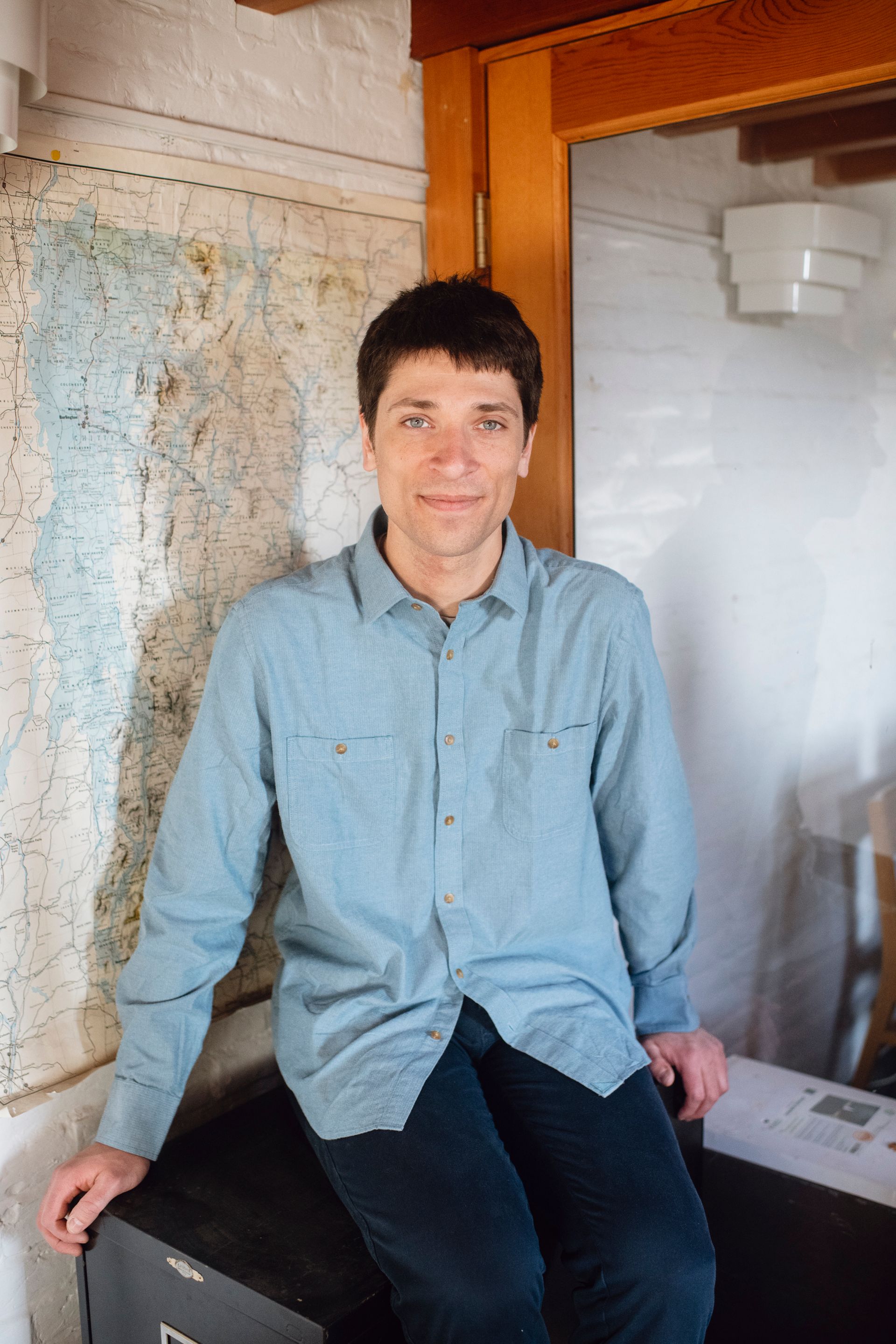 Man seated indoors beside a wall map, wearing a light blue shirt and dark pants, near a wooden doorway