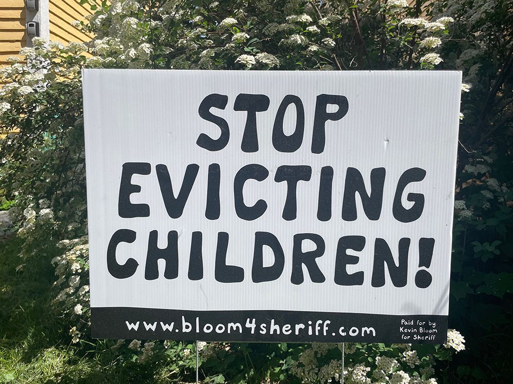 White protest sign reads “STOP EVICTING CHILDREN!” with bloom4sheriff.com at the bottom.