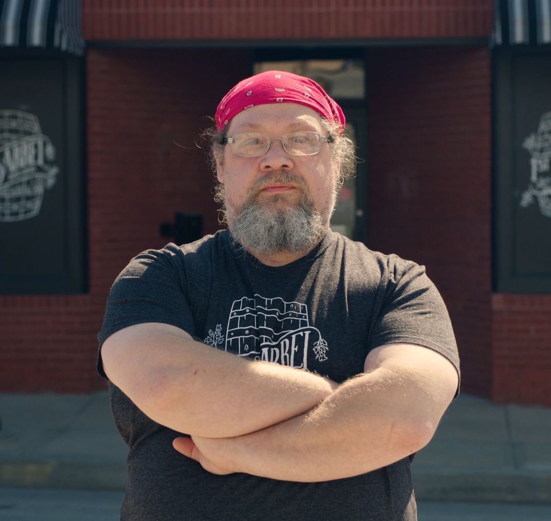 A man with a beard and glasses is standing with his arms crossed in front of a brick building.