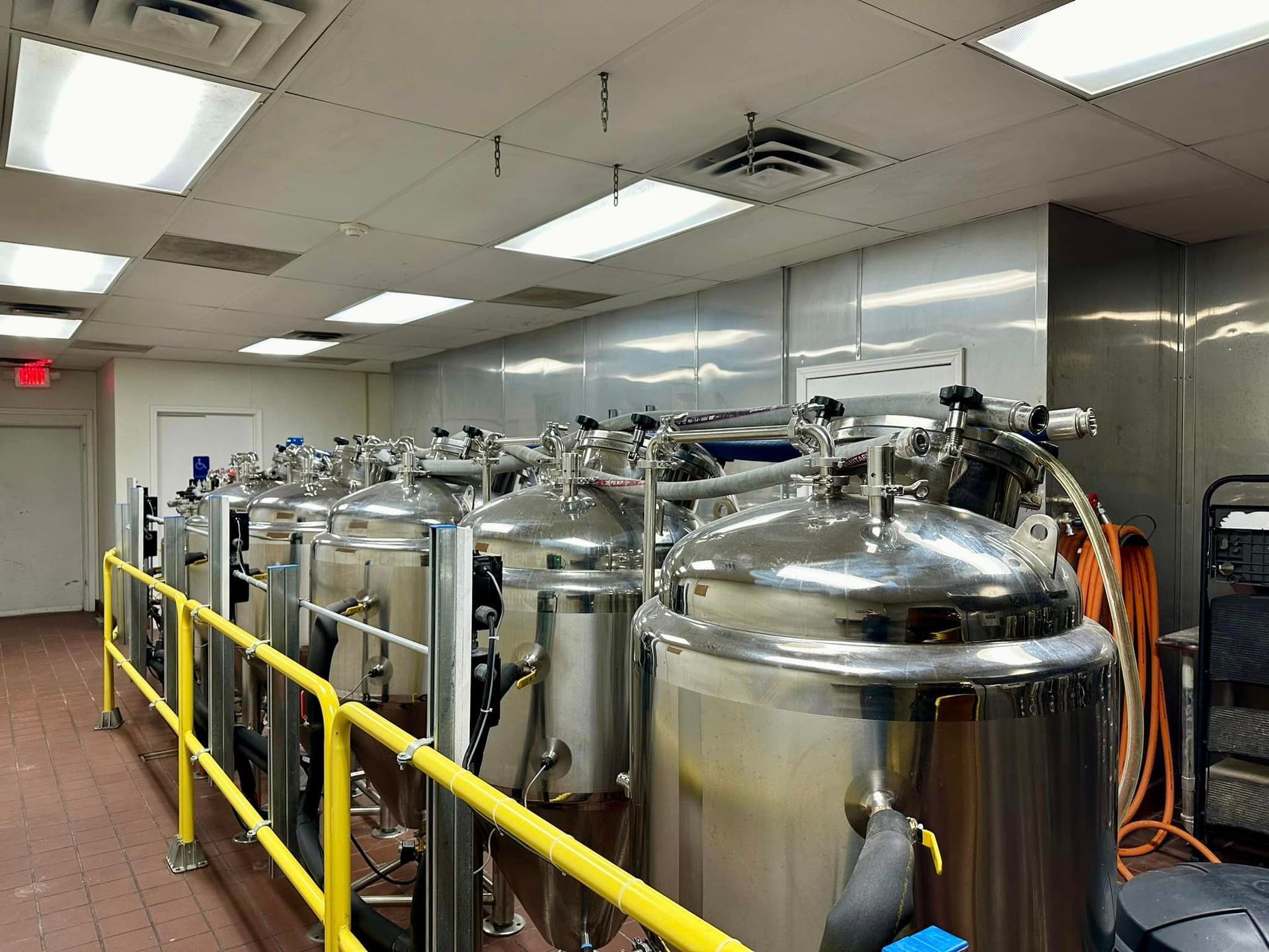 A row of stainless steel tanks are lined up in a room with a yellow railing.