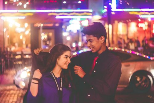 Young couple smiles at each other on a night street, neon lights in the background.