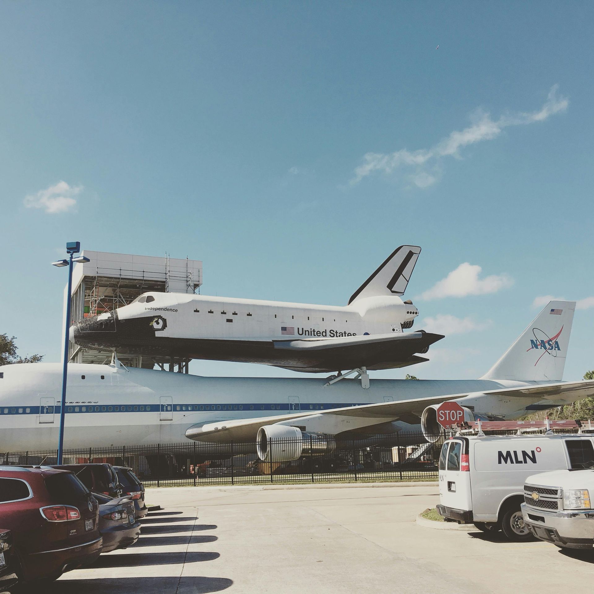 Space Shuttle atop NASA Boeing 747; outdoors, cars parked nearby, stop sign, blue sky.