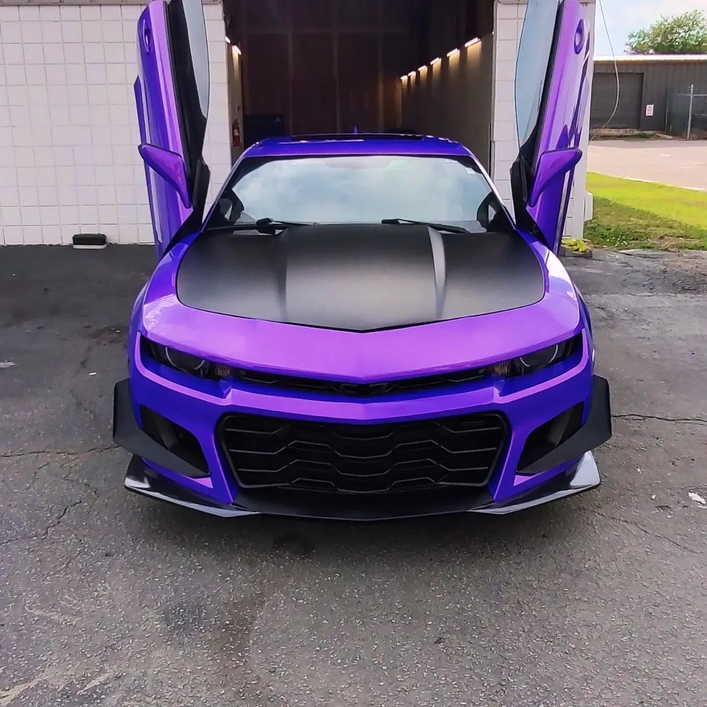 Purple sports car with black hood and doors open, parked in front of a building.