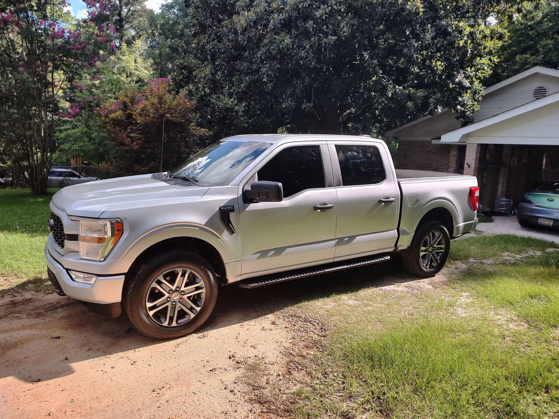 Silver Ford F-150 pickup truck parked on a driveway with a house and trees in the background.