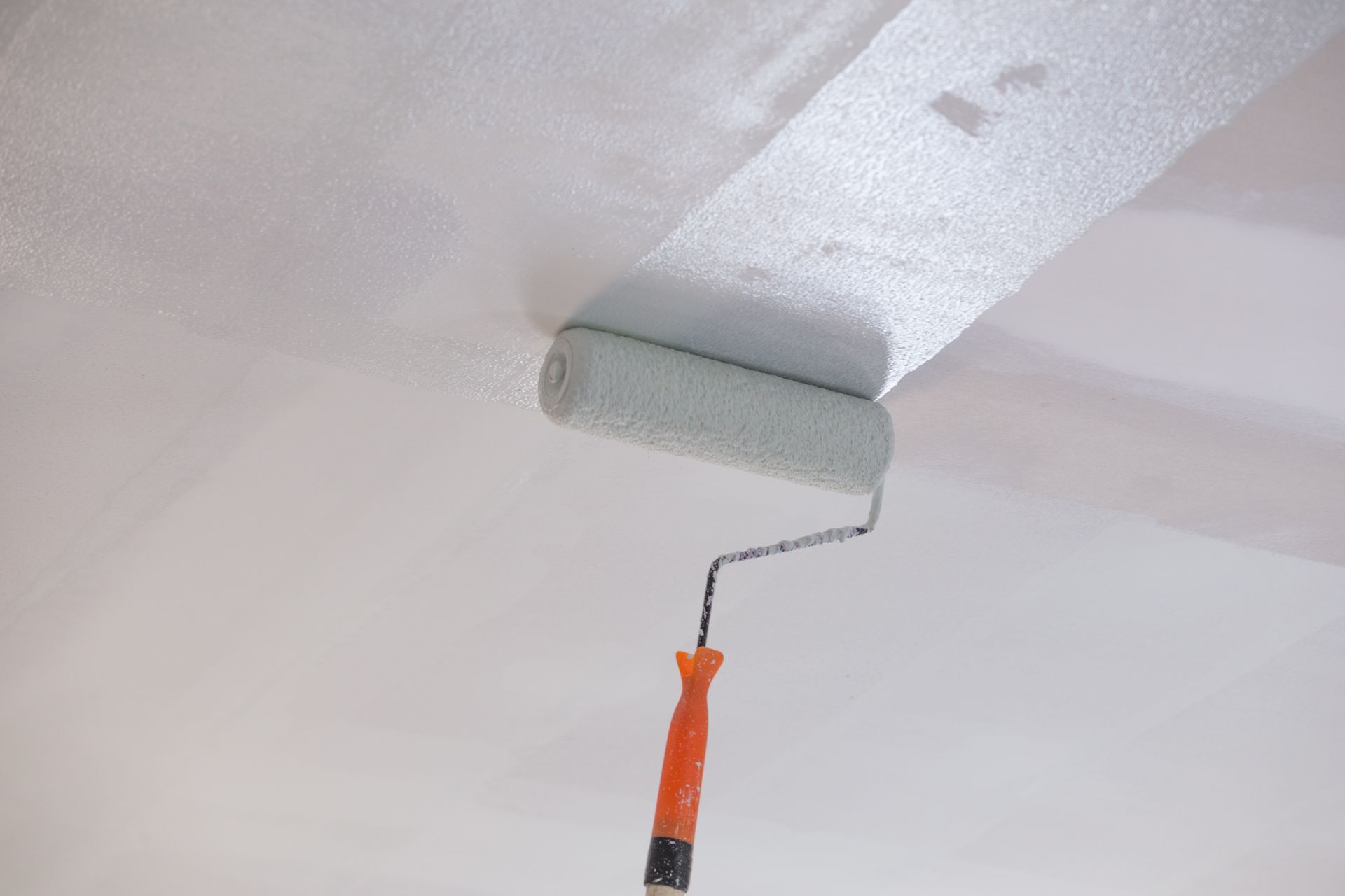 A paint roller applies a layer of white paint to a ceiling surface.