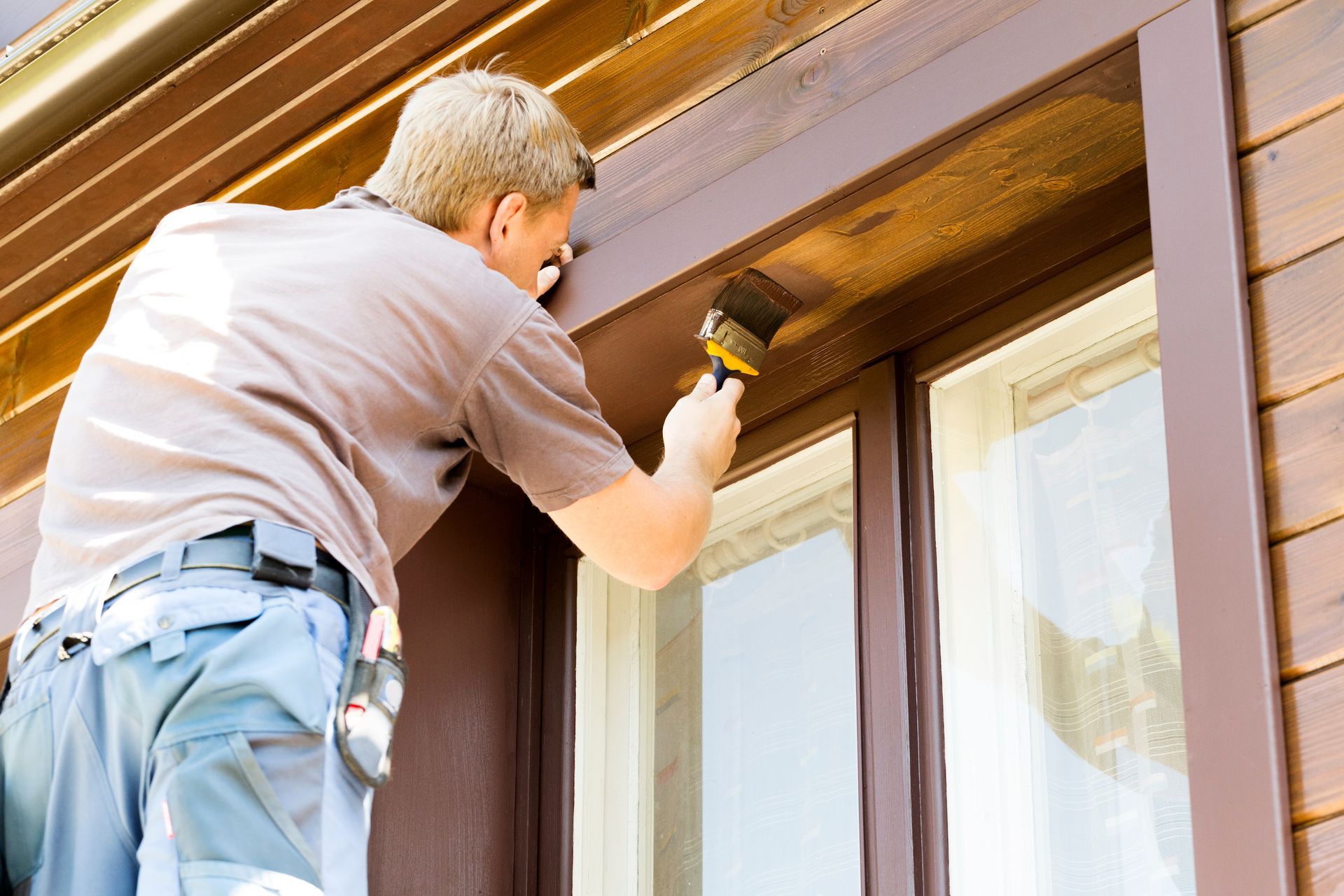 A person in a brown shirt and light work pants paints the wooden trim above a window on a house.