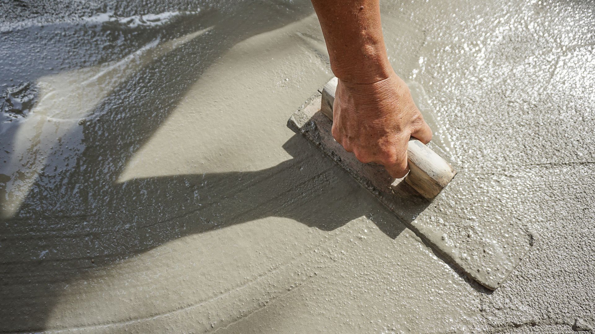 A hand uses a wooden float to smooth a wet layer of gray concrete.