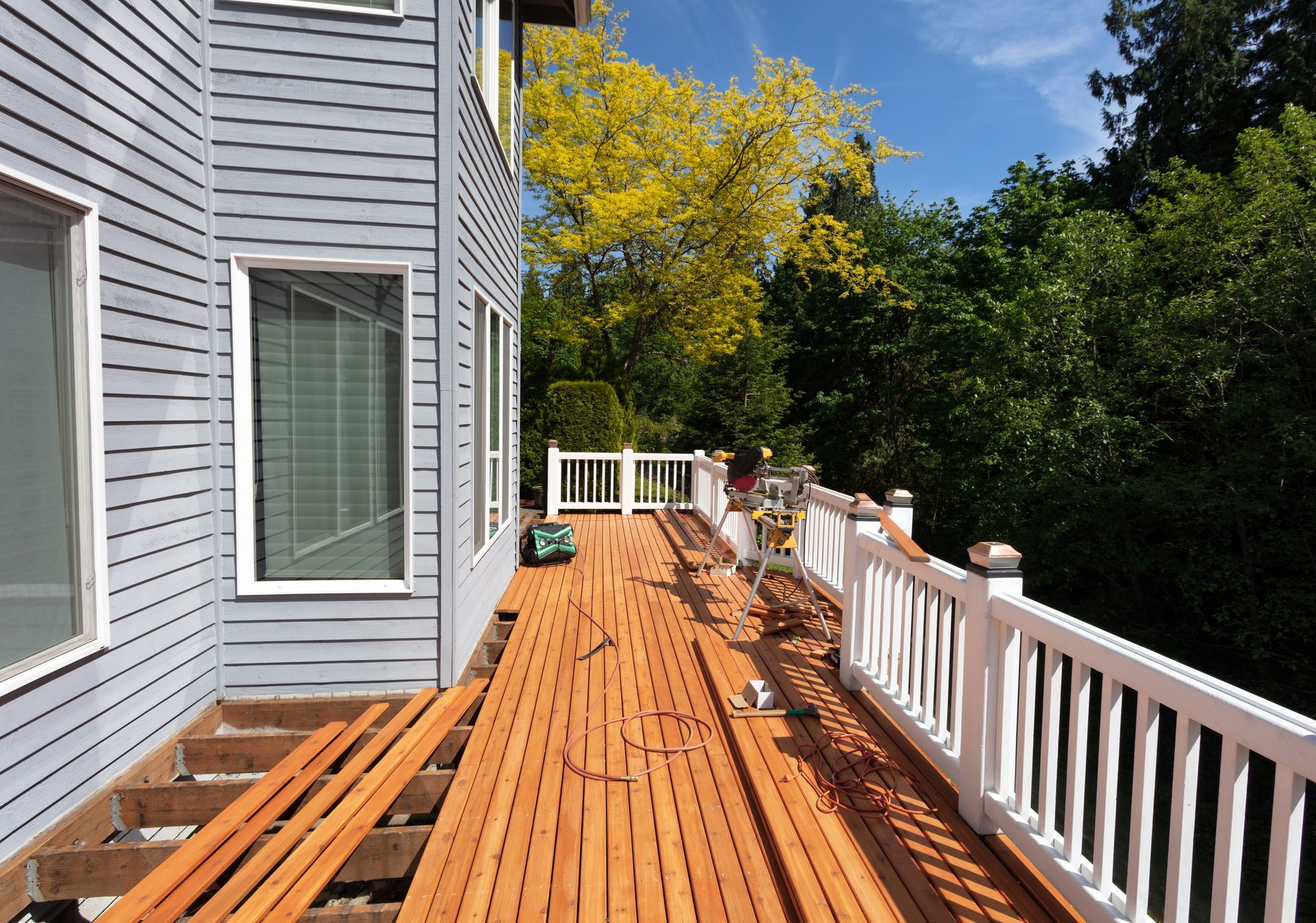 A partially finished wooden deck attached to a gray house, featuring white railings and lush trees in the background.