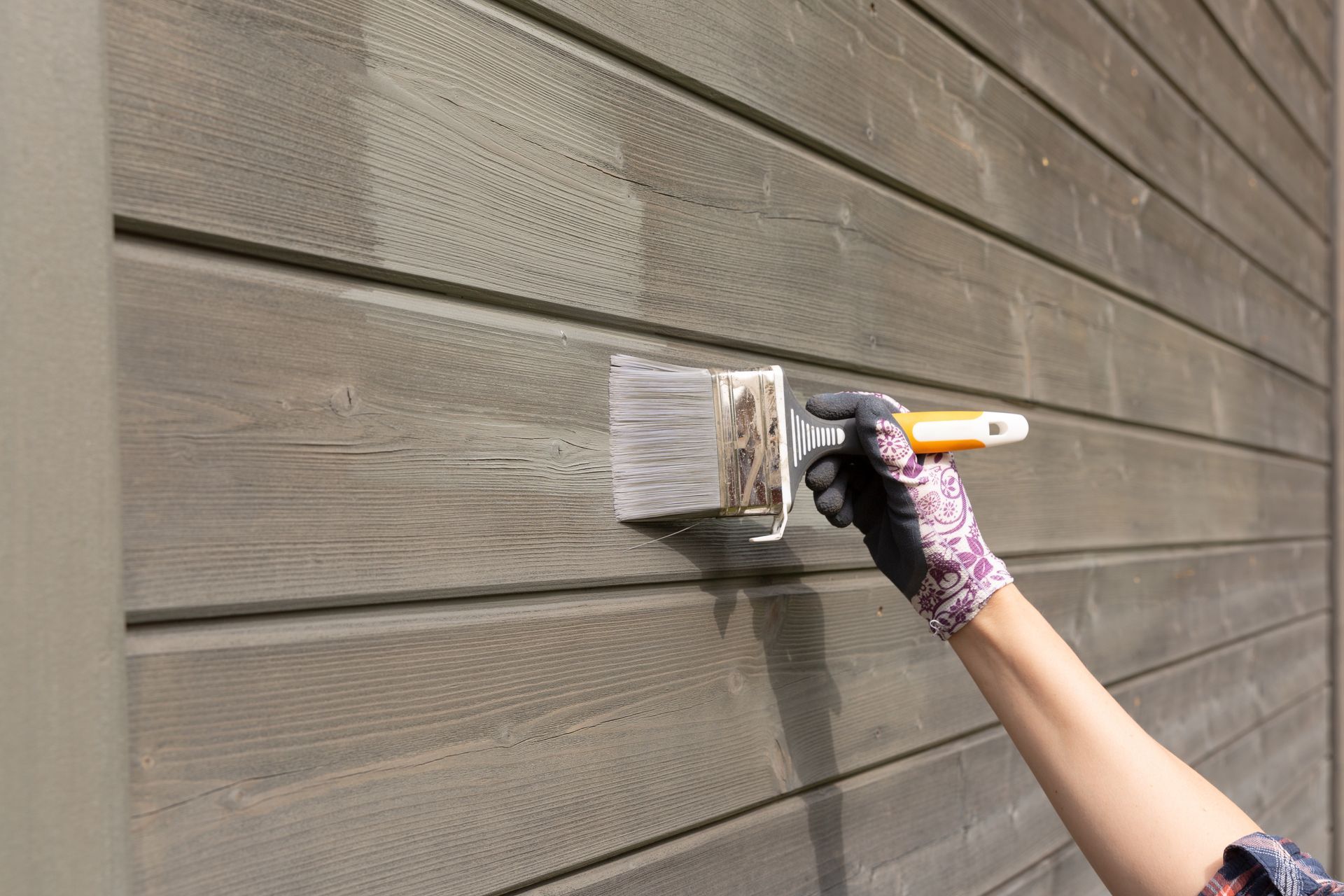 A hand in a patterned glove uses a wide brush to apply stain to horizontal wooden siding.
