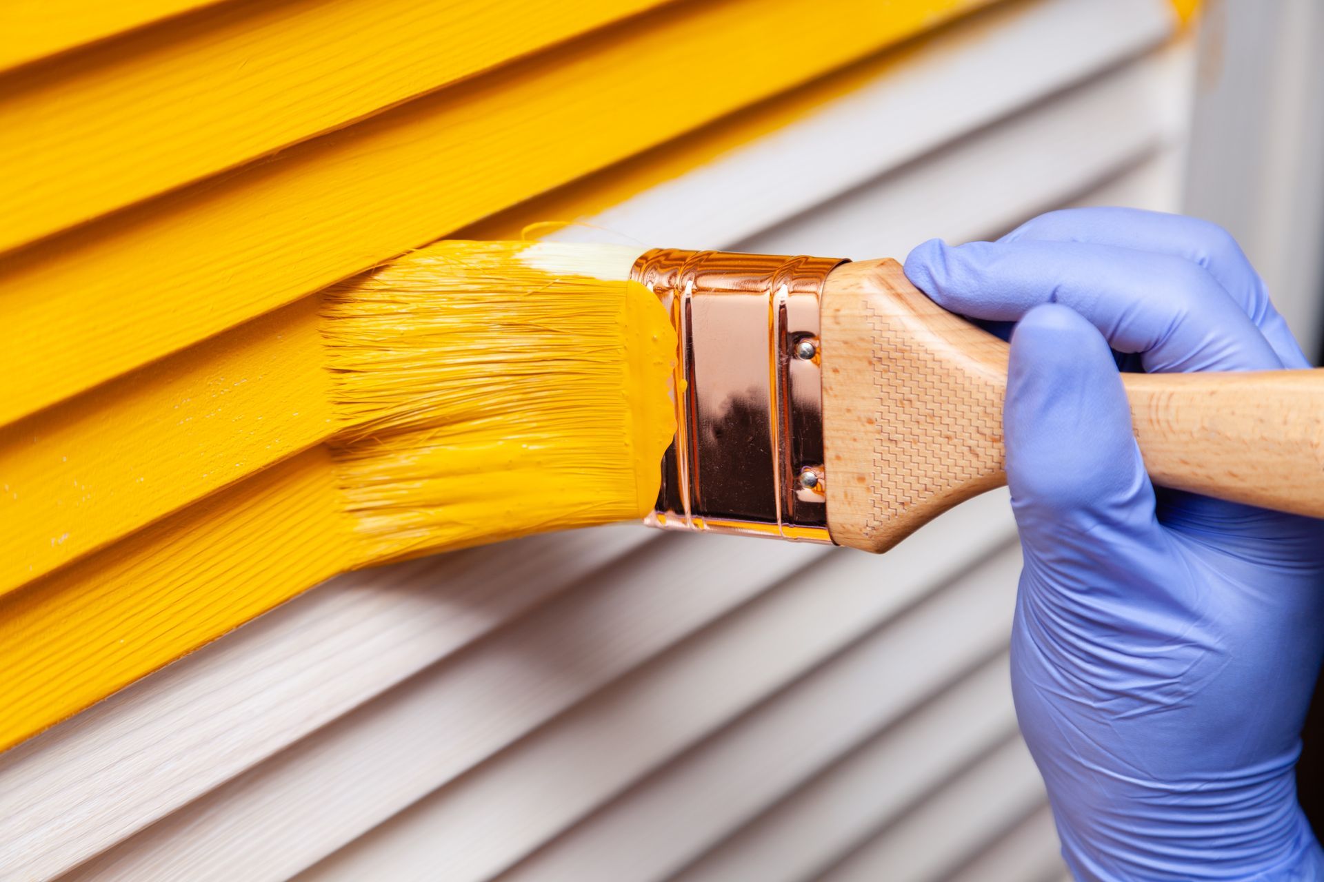 A hand wearing a blue nitrile glove uses a brush to paint white louvered blinds with bright yellow paint.