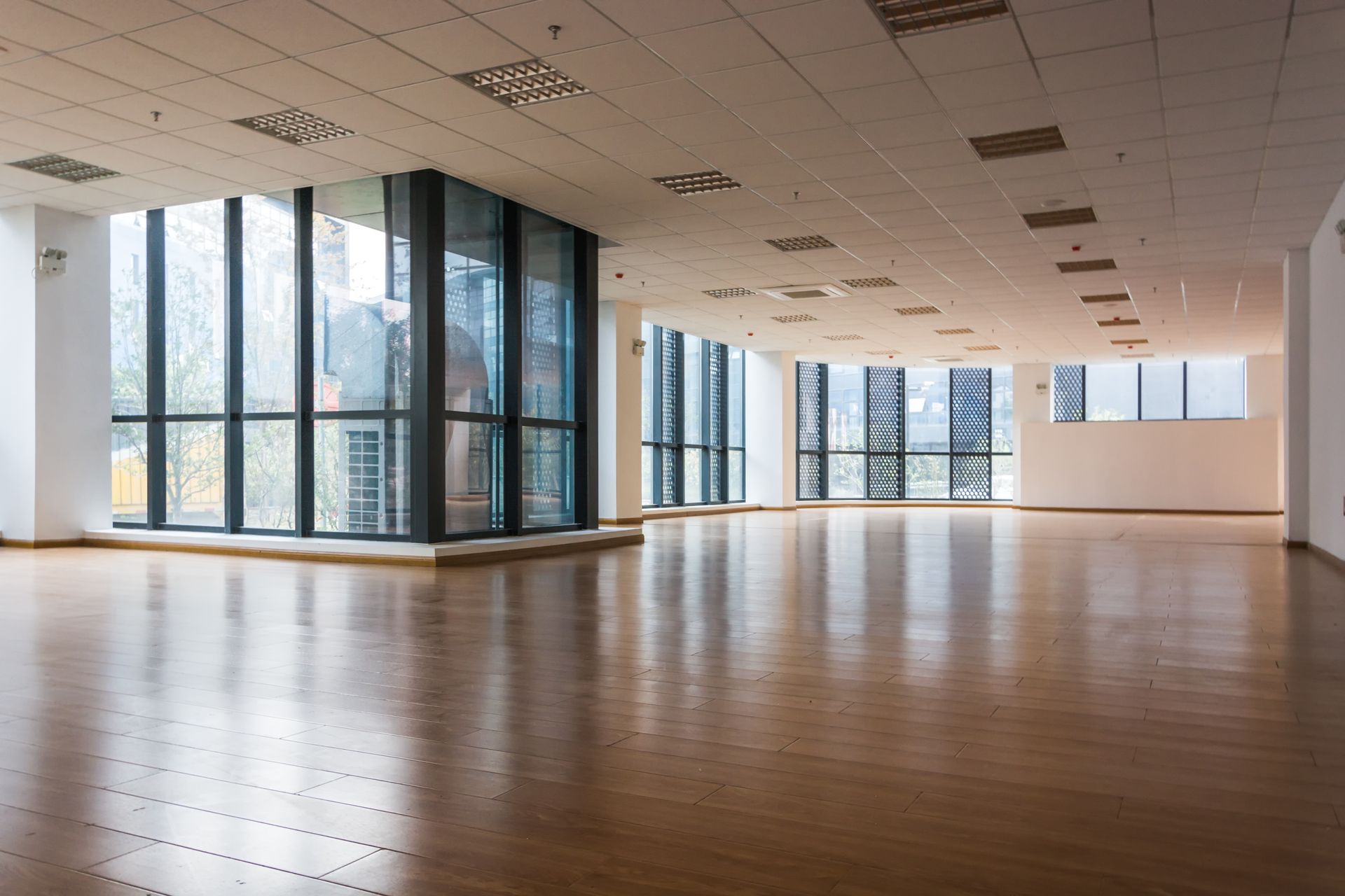 A bright, empty open-plan office with polished wood floors, high ceilings, and floor-to-ceiling windows.