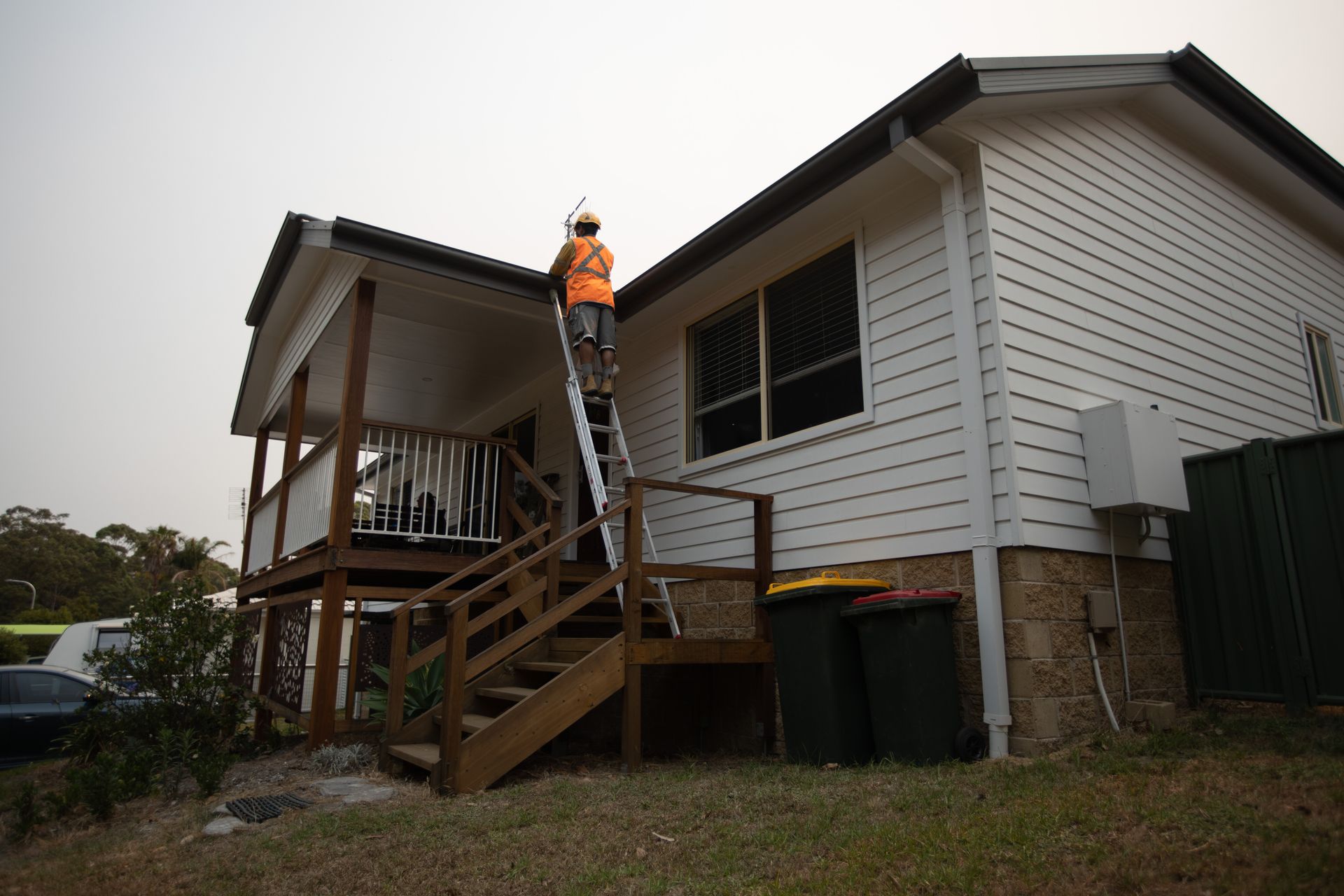 A person in an orange high-visibility vest climbs a ladder against the side of a white house to inspect the roof.