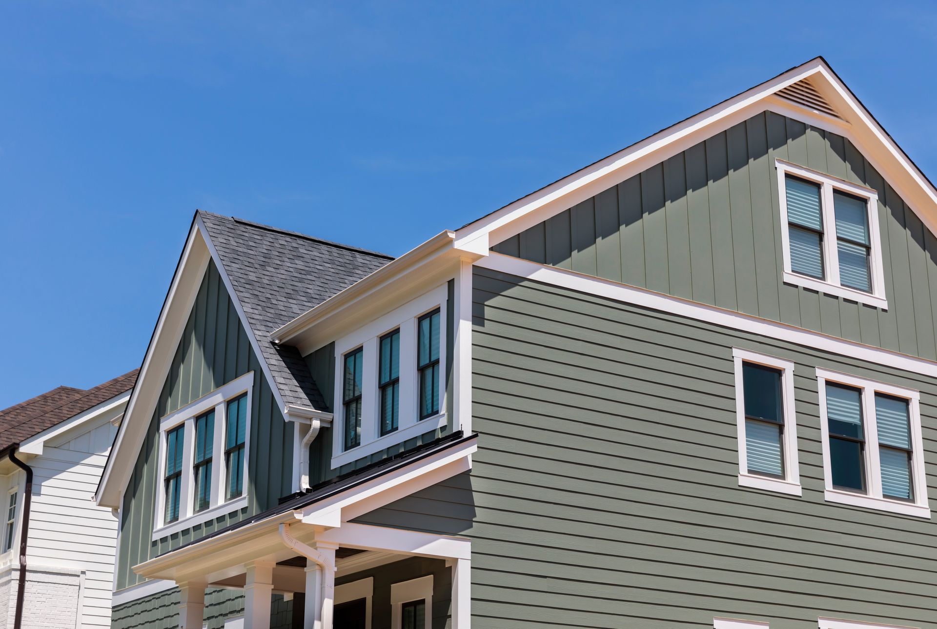 A large, multi-story house with green siding, white trim, and dark shingled roofs under a clear blue sky.