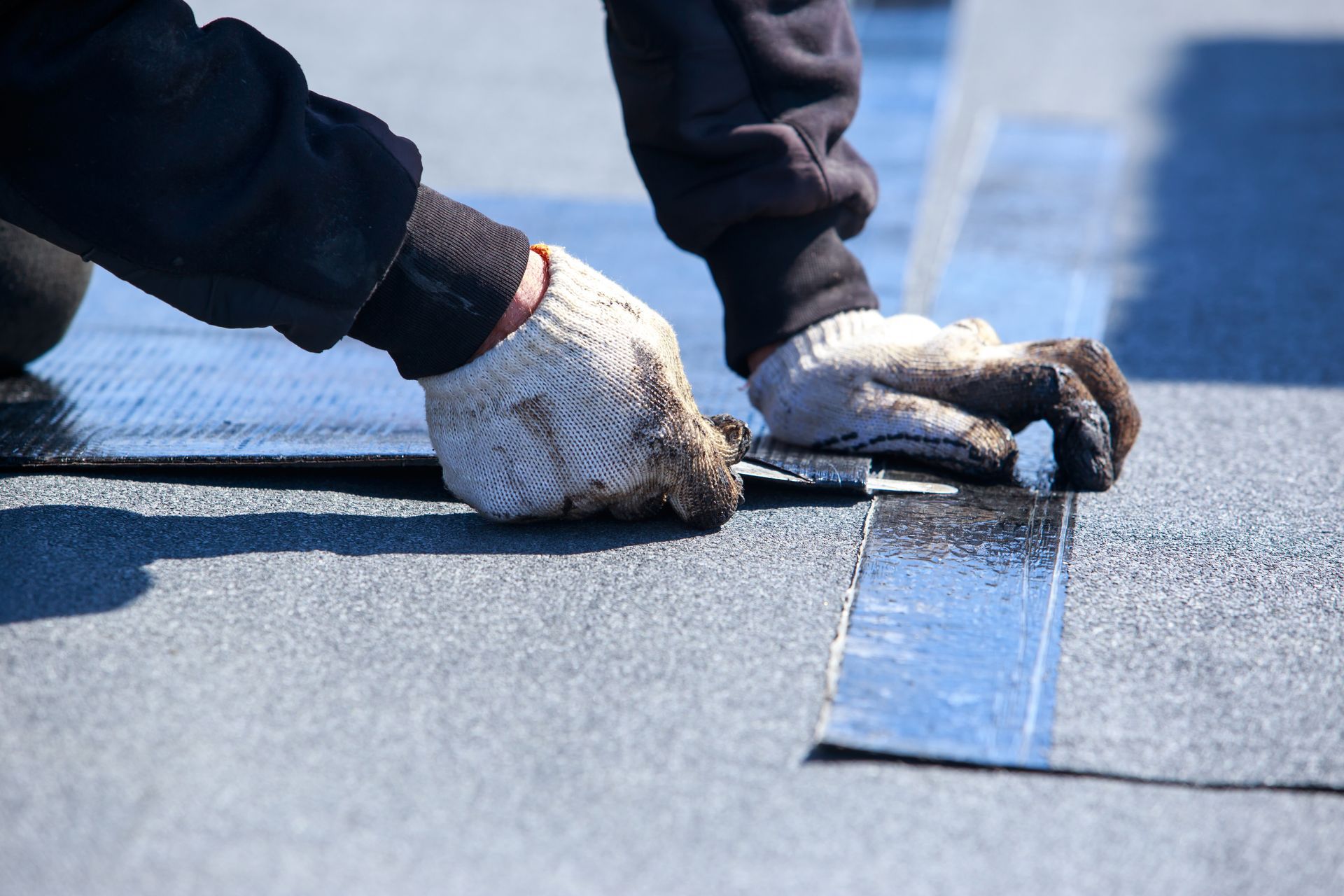 A worker wearing work gloves applies a strip of adhesive roofing membrane to a flat roof surface.