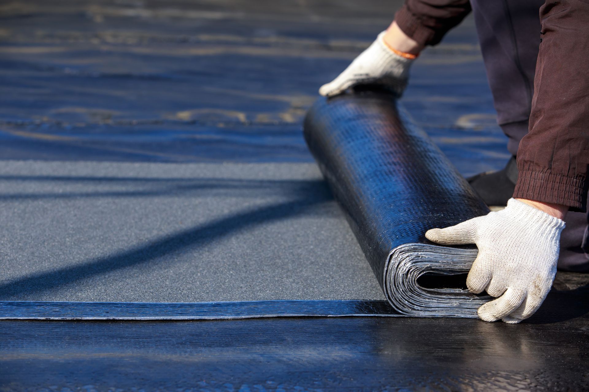 A worker in white work gloves unrolls a strip of black bitumen roofing material onto a dark, prepared roof surface.