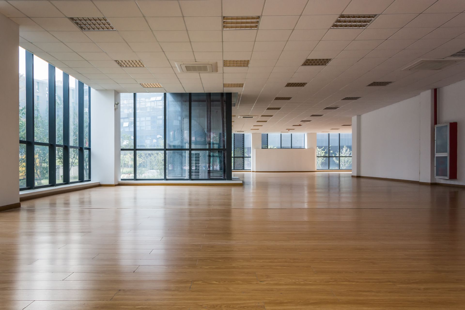 An empty, modern office space with floor-to-ceiling windows, light wood flooring, and a drop ceiling.