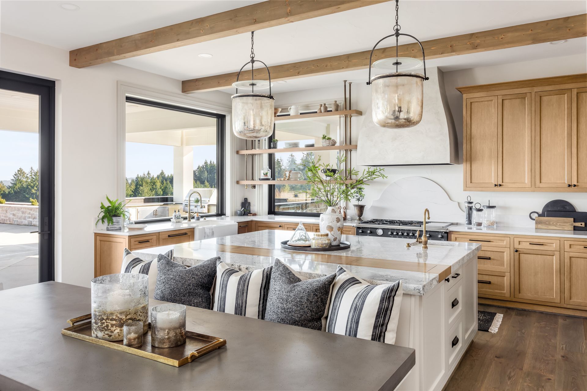 A bright, modern kitchen with light wood cabinets, a large island with patterned stools, and rustic ceiling beams.