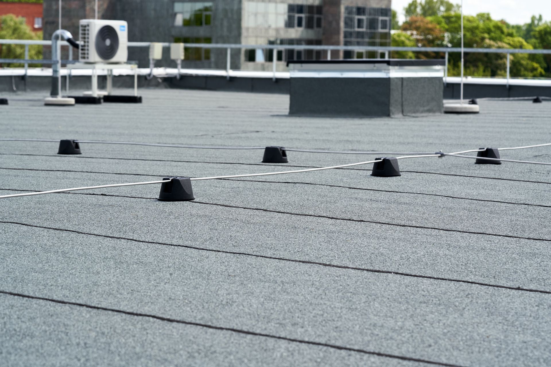 A flat, gray, asphalt-covered roof featuring small black mounting fixtures and an HVAC unit under a cloudy sky.