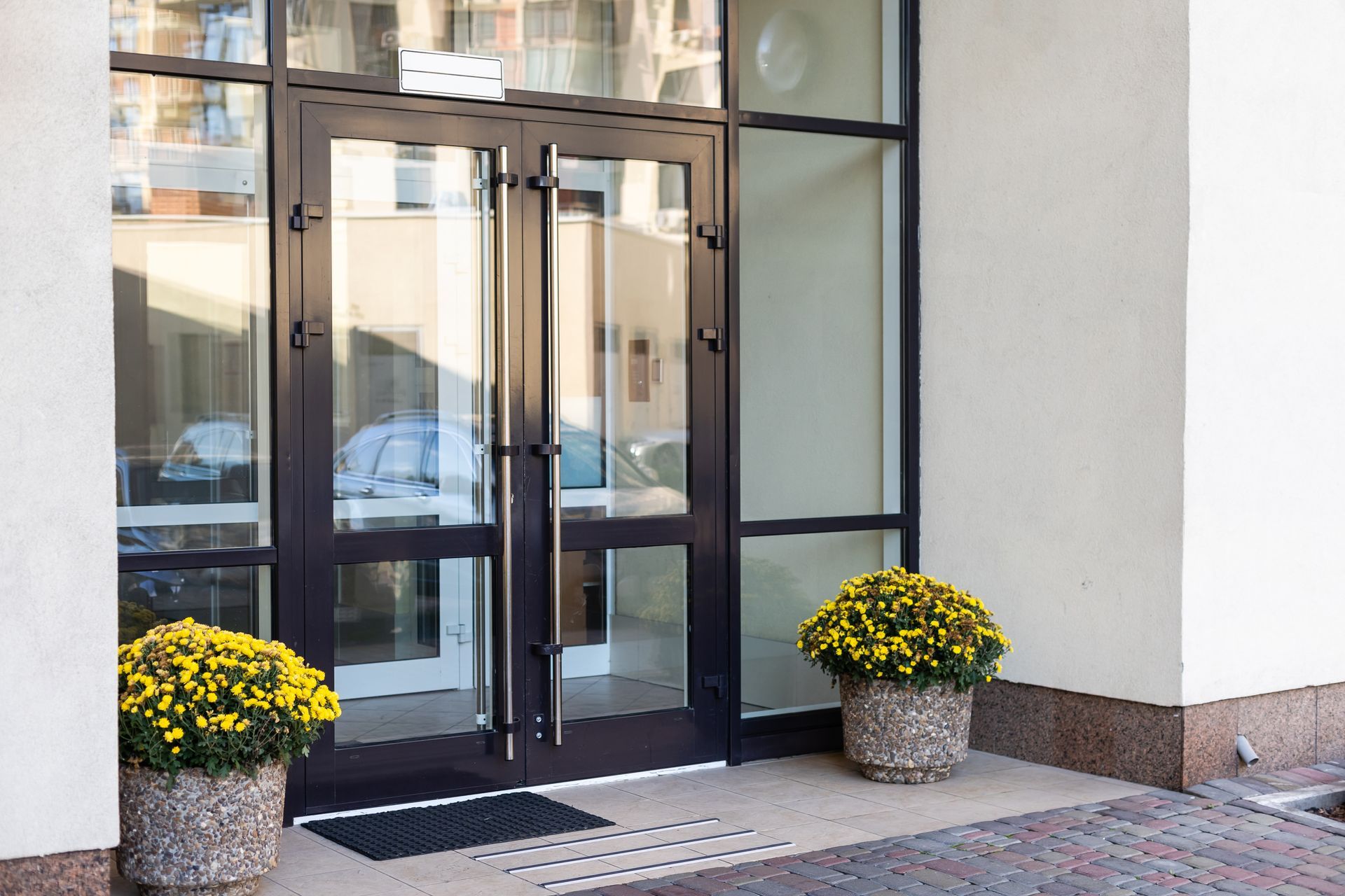 A glass double door entrance with dark metal frames, flanked by two stone planters filled with yellow chrysanthemums.