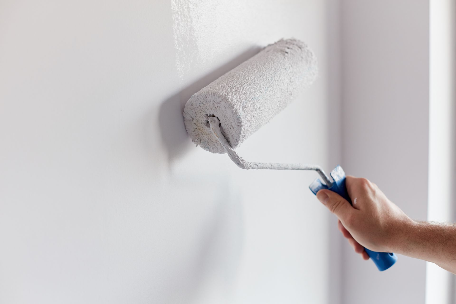 A hand holds a paint roller, applying a coat of white paint to a smooth, light-colored wall.