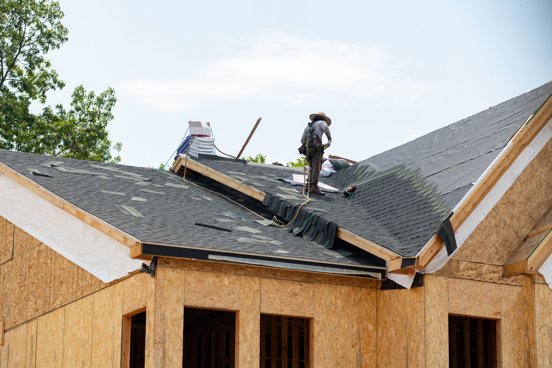A worker installing shingles on the wooden frame of a house roof under a clear sky.