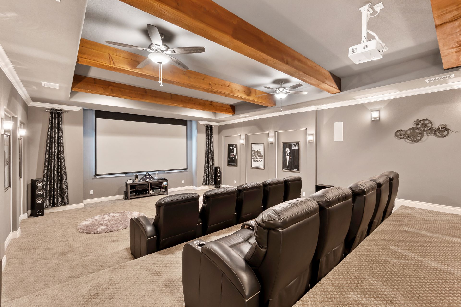 A home theater with rows of black leather reclining chairs facing a large projection screen, beneath exposed wood beams.