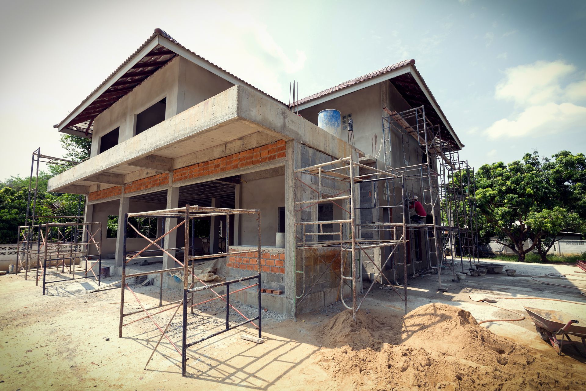 A house under construction with a metal scaffold frame in the foreground, concrete walls, and a tiled roof.